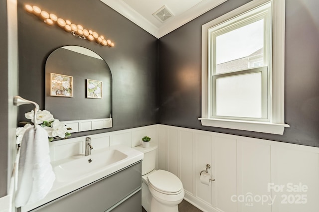 half bath with crown molding, vanity, toilet, a wainscoted wall, and visible vents