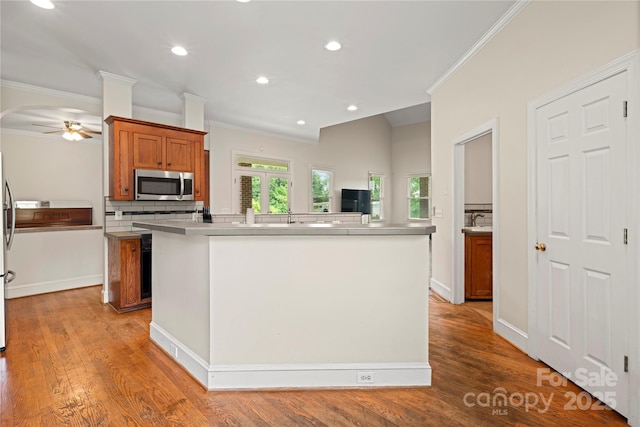 kitchen with stainless steel microwave, crown molding, a center island with sink, brown cabinetry, and a ceiling fan
