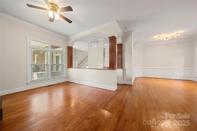 empty room featuring wood finished floors, ornamental molding, ceiling fan, arched walkways, and ornate columns