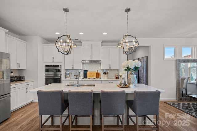 kitchen with a chandelier, appliances with stainless steel finishes, a sink, under cabinet range hood, and backsplash