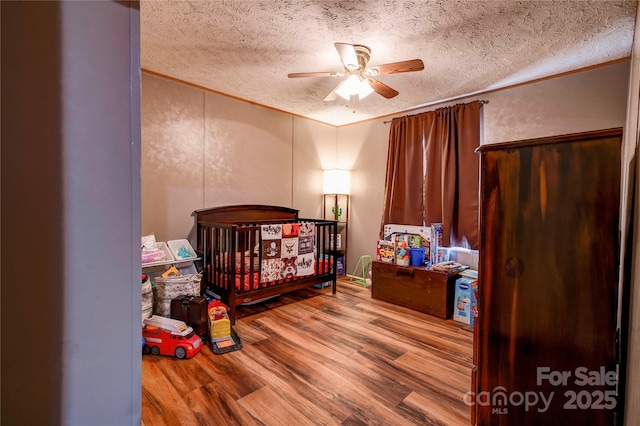 bedroom with wood finished floors, a crib, ornamental molding, a ceiling fan, and a textured ceiling