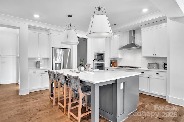 kitchen featuring a kitchen island with sink, light countertops, wall chimney exhaust hood, tasteful backsplash, and appliances with stainless steel finishes