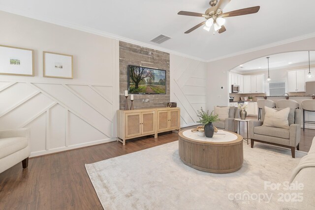 living room featuring ceiling fan, visible vents, a decorative wall, ornamental molding, and wood finished floors