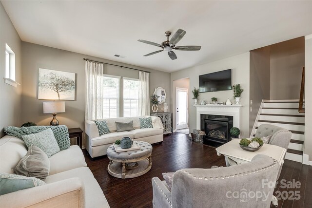 living area featuring a ceiling fan, dark wood-style flooring, visible vents, a glass covered fireplace, and stairs