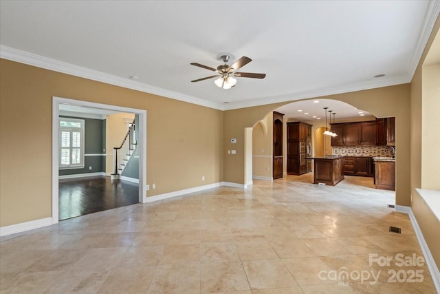 unfurnished living room with arched walkways, crown molding, a ceiling fan, baseboards, and a sink