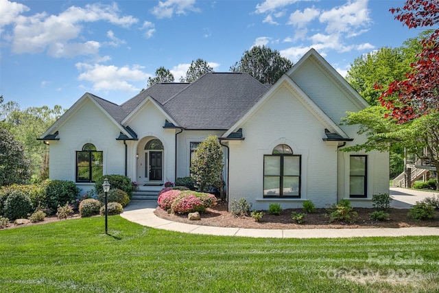 french country home featuring brick siding, a front lawn, and roof with shingles
