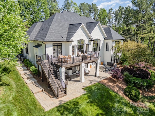 rear view of property with a wooden deck, a patio area, a yard, a shingled roof, and stairs