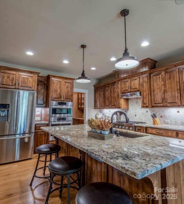kitchen featuring appliances with stainless steel finishes, light stone counters, decorative backsplash, brown cabinetry, and a sink