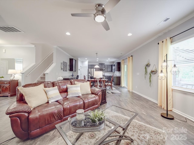 living room with ceiling fan, light wood-type flooring, visible vents, ornamental molding, and stairway