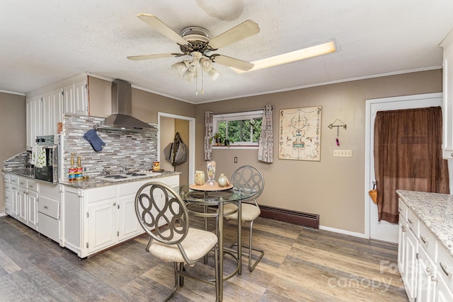 kitchen featuring wood finished floors, a ceiling fan, wall chimney exhaust hood, tasteful backsplash, and white cabinetry