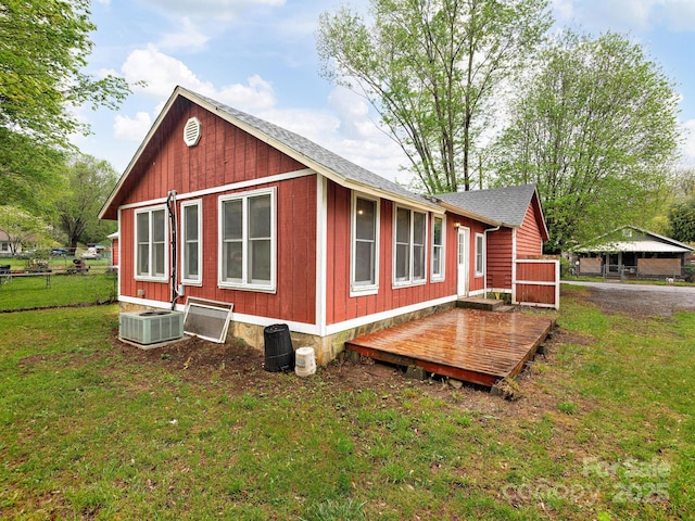 view of side of home with fence, a deck, cooling unit, roof with shingles, and a lawn