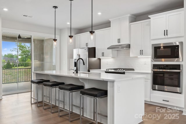 kitchen featuring a kitchen island with sink, decorative backsplash, stainless steel appliances, under cabinet range hood, and wood finished floors