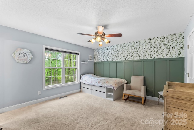 carpeted bedroom featuring wainscoting, a textured ceiling, a ceiling fan, wallpapered walls, and visible vents