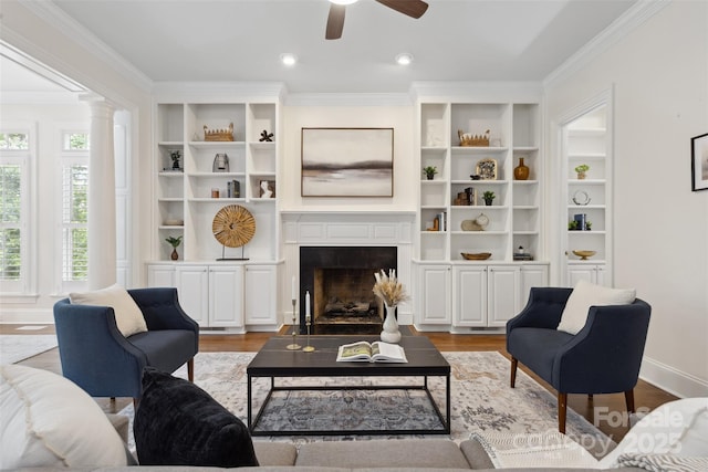 living area featuring a ceiling fan, decorative columns, a fireplace, wood finished floors, and ornamental molding