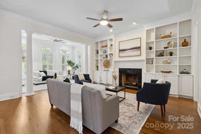 living area featuring ceiling fan, crown molding, a fireplace, wood finished floors, and ornate columns