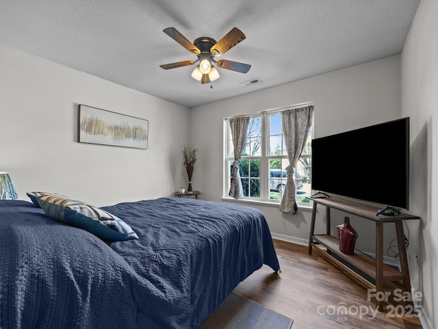 bedroom with ceiling fan, a textured ceiling, baseboards, wood finished floors, and visible vents