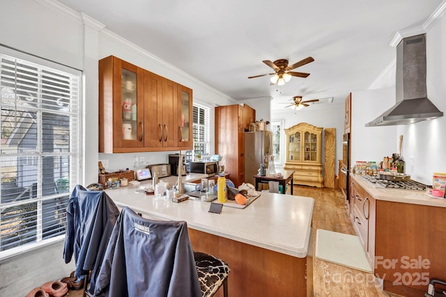 kitchen with wall chimney range hood, ceiling fan, brown cabinetry, light countertops, and a peninsula