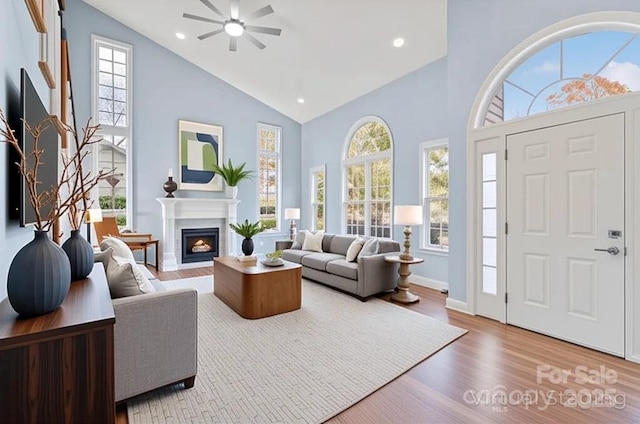 living area with a warm lit fireplace, high vaulted ceiling, a ceiling fan, recessed lighting, and wood finished floors