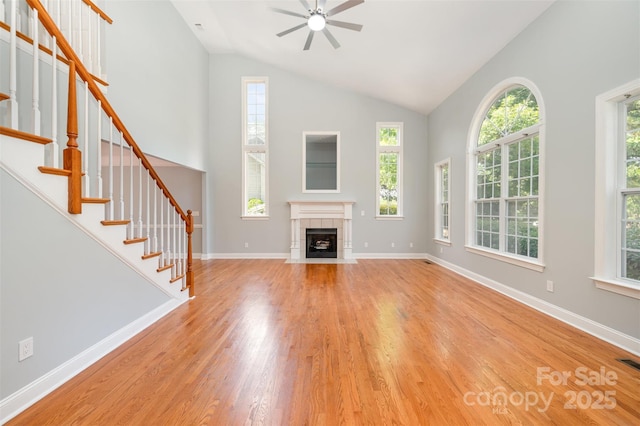 unfurnished living room with wood finished floors, stairway, high vaulted ceiling, a tile fireplace, and a ceiling fan
