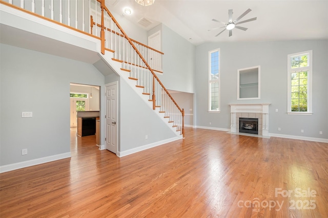 unfurnished living room with a fireplace, ceiling fan, high vaulted ceiling, stairway, and light wood-type flooring