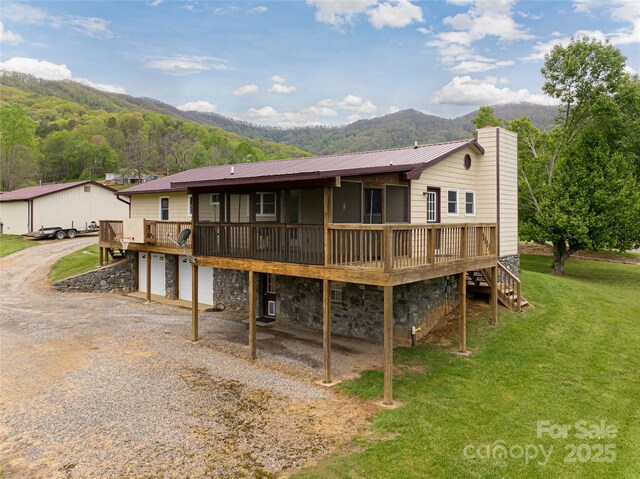 back of house with an attached garage, a deck with mountain view, a yard, driveway, and metal roof