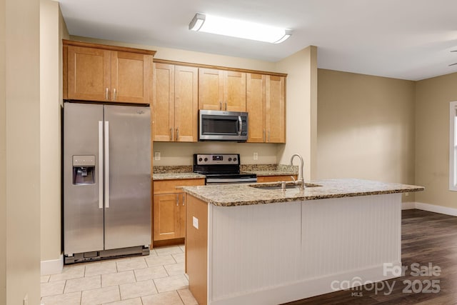kitchen featuring appliances with stainless steel finishes, light stone counters, a sink, a kitchen island with sink, and baseboards