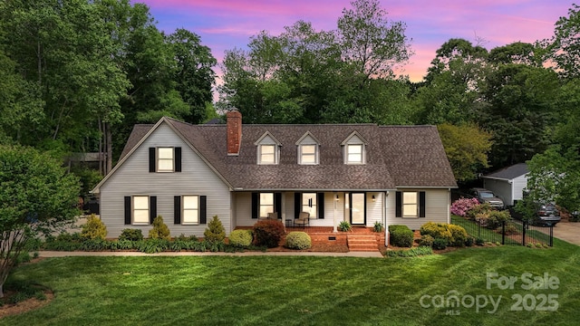 cape cod house with roof with shingles, covered porch, a yard, and a chimney