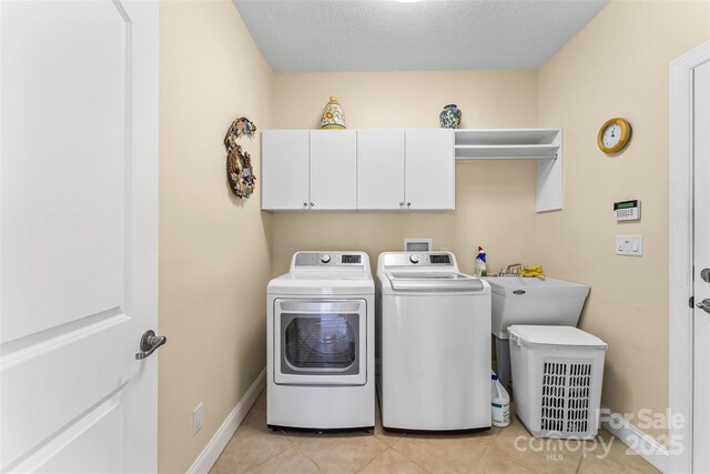 clothes washing area with a textured ceiling, washing machine and dryer, cabinet space, baseboards, and light tile patterned flooring