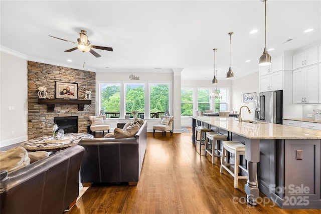 living area with a ceiling fan, crown molding, a stone fireplace, dark wood-style floors, and recessed lighting