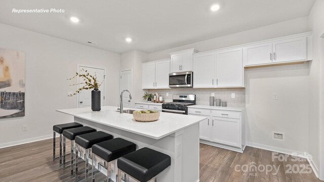 kitchen with a kitchen island with sink, appliances with stainless steel finishes, a sink, white cabinetry, and dark wood-type flooring