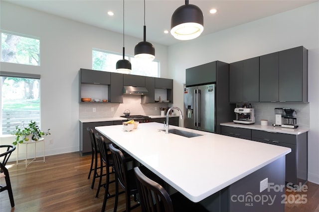 kitchen with a sink, light countertops, high end refrigerator, dark wood-type flooring, and under cabinet range hood