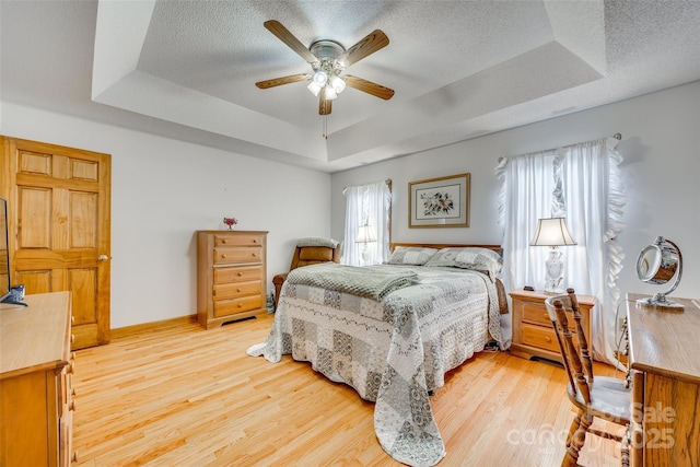 bedroom with ceiling fan, a raised ceiling, light wood-style flooring, baseboards, and a textured ceiling