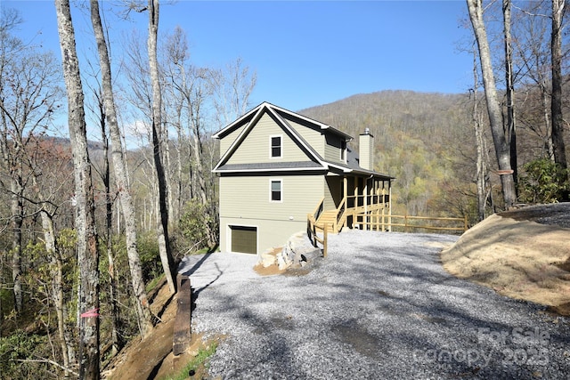 view of side of home with fence, gravel driveway, a chimney, an attached garage, and a forest view