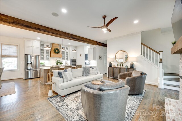 living area featuring a ceiling fan, stairs, light wood finished floors, and beam ceiling