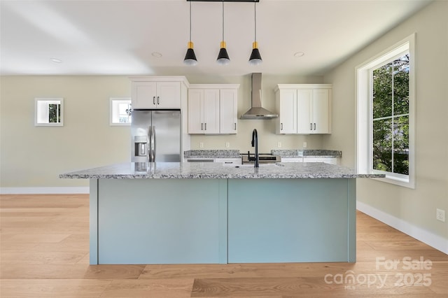 kitchen with light stone countertops, an island with sink, wall chimney range hood, stainless steel fridge, and white cabinets