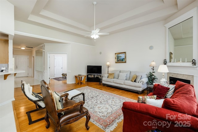living room with light wood finished floors, a fireplace, a ceiling fan, baseboards, and a tray ceiling