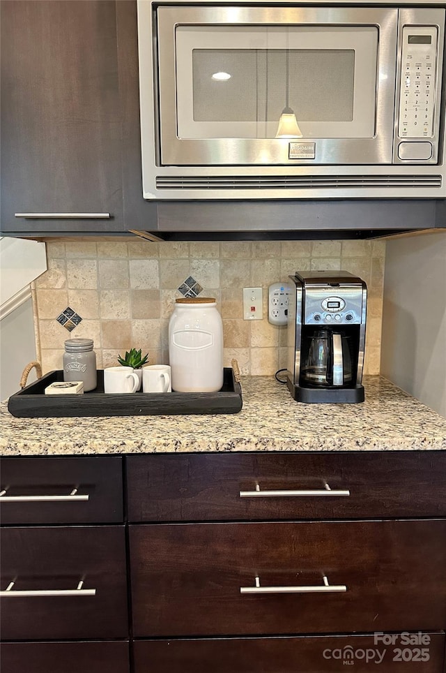 details with backsplash, stainless steel microwave, dark brown cabinetry, and light stone countertops