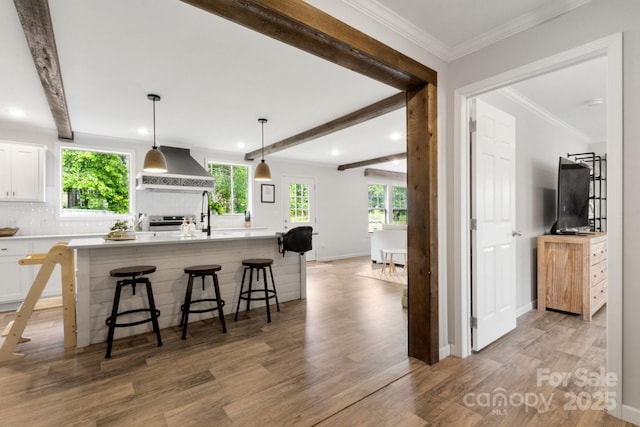 kitchen with wall chimney range hood, an island with sink, white cabinetry, light wood-type flooring, and a kitchen bar