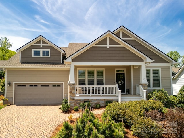 craftsman-style house with a porch, roof with shingles, decorative driveway, an attached garage, and stone siding