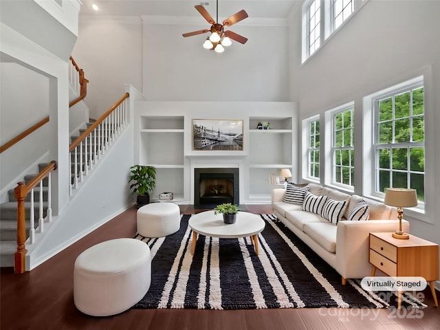 living area featuring a ceiling fan, a towering ceiling, a fireplace with flush hearth, built in features, and stairs