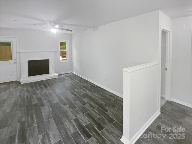 unfurnished living room featuring a brick fireplace, a ceiling fan, baseboards, and dark wood-type flooring