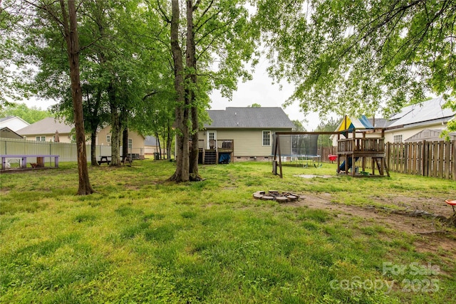 view of yard with a trampoline, a wooden deck, a fenced backyard, a playground, and an outdoor fire pit