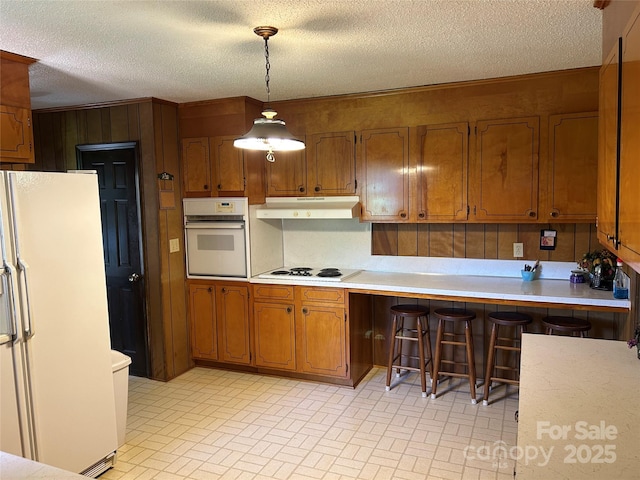 kitchen featuring brown cabinetry, white appliances, under cabinet range hood, light countertops, and brick patterned floor