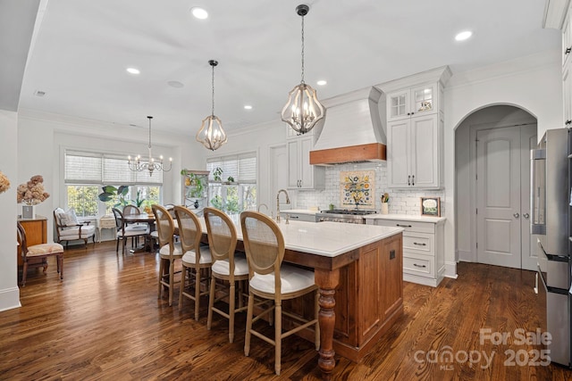 kitchen featuring tasteful backsplash, a chandelier, custom range hood, dark wood-style floors, and a center island with sink