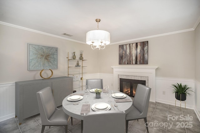 tiled dining room with a chandelier, ornamental molding, a brick fireplace, and a wainscoted wall