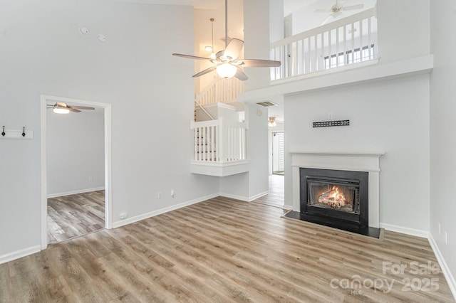 unfurnished living room with wood finished floors, baseboards, ceiling fan, a high ceiling, and a fireplace with flush hearth