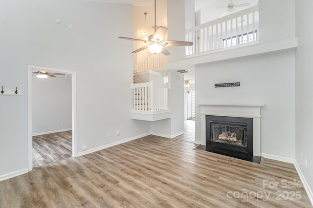 unfurnished living room featuring ceiling fan, baseboards, a fireplace with flush hearth, wood finished floors, and a towering ceiling