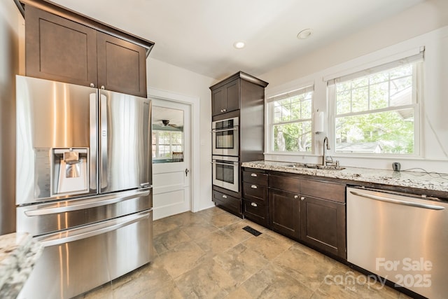 kitchen with stone finish floor, appliances with stainless steel finishes, dark brown cabinets, a sink, and light stone counters