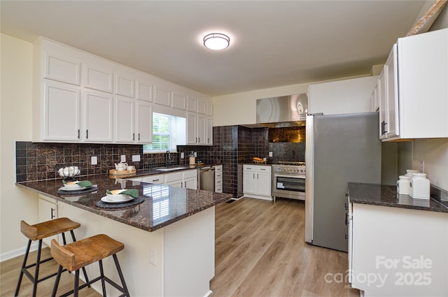 kitchen featuring a sink, appliances with stainless steel finishes, wall chimney exhaust hood, a peninsula, and light wood-style floors
