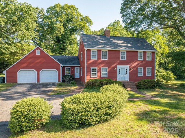 colonial inspired home with a shingled roof, an attached garage, a front lawn, a chimney, and driveway